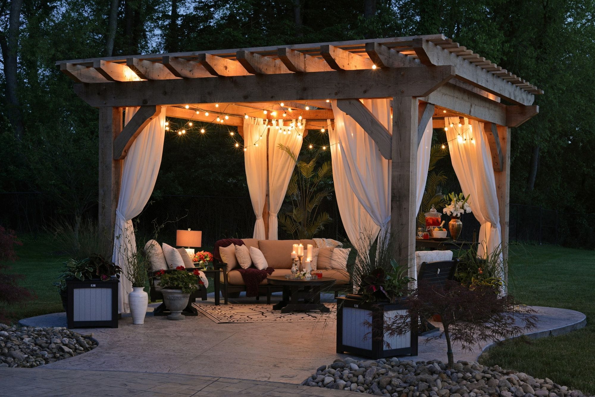 Pergola Salonu — Ahşap pergola, tül perdeler ve sıcak ışıkla bahçede bir açık hava salonu.